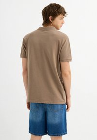 Camisa polo de algodón marrón con mangas cortas, que presenta un cuello clásico y un corte ligeramente relajado, combinada con shorts de mezclilla azul claro.