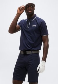 Άνδρας ντυμένος με ναυτικό μπλε γκολφ ένδυμα, που διορθώνει λευκό καπέλο με το αριστερό χέρι, φορώντας λευκό γάντι γκολφ στο δεξί χέρι, σε απλό φόντο.