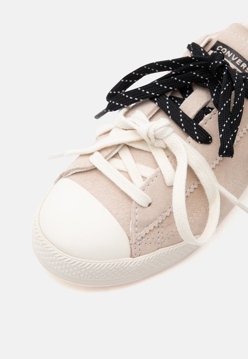 Beiger Sneaker mit weißer Gummispitze, schwarz-weiß gepunkteten Schnürsenkeln über weißen Schnürsenkeln, die zu einer Schleife gebunden sind, und sichtbarem "Converse"-Etikett auf der Zunge.