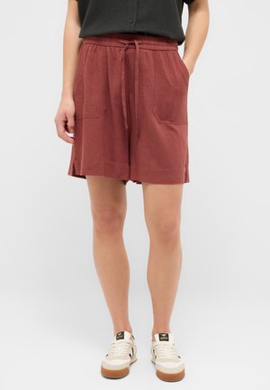 Person wearing rust-colored shorts with front pockets, a black top, and white sneakers with gum soles, standing against a plain background.