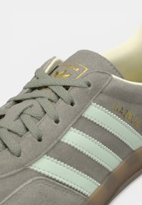 Γκρι σουέτ παπούτσι με ριγέ σε μέντα και χρυσές λεπτομέρειες λογότυπου. Στρογγυλερό μπροστινό μέρος, υφασμένα κορδόνια και ελαφριά ελαστική σόλα ορατή.