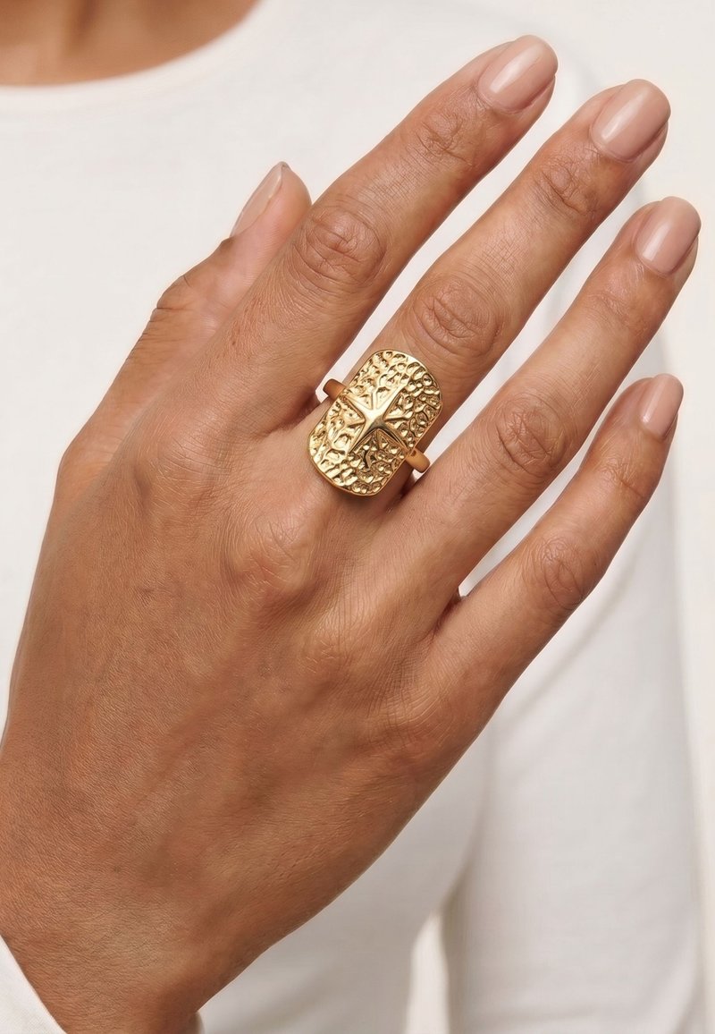 Main avec une peau brun moyen portant une grande bague en or texturée avec une croix, présentée sur un vêtement blanc à manches longues.