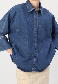 Camicia di denim con una sfumatura blu, caratterizzata da intagli a forma di cuore. Chiusura frontale con bottoni e vestibilità rilassata con maniche ampie. Tessuto stondato.