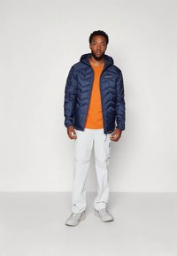 Giacca a piumino navy con motivo a chevron, t-shirt arancione, pantaloni cargo bianchi e sneakers grigio chiaro. Caratterizzata da cappuccio e chiusura con cerniera.