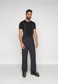 T-shirt preto combinado com calças escuras, impermeáveis, com alças ajustáveis e bolsos. Complementa com botas de esqui pretas. Design minimalista.