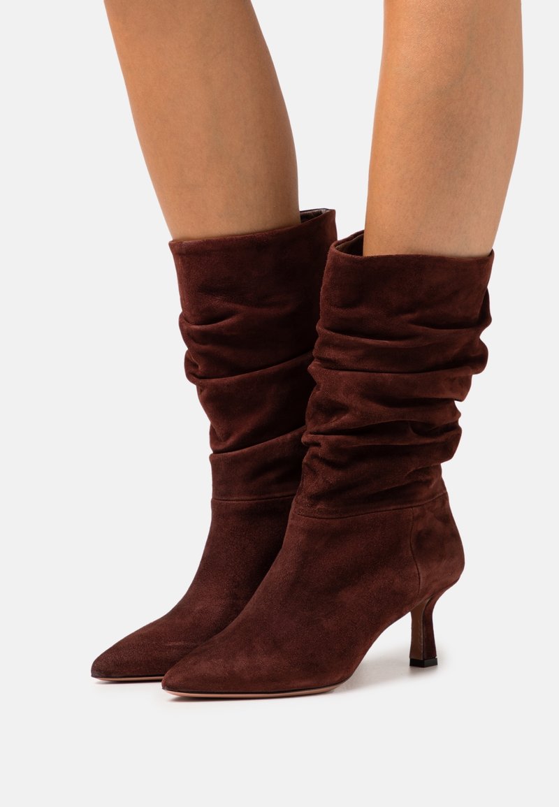 Bottes décontractées en daim marron foncé montant jusqu'au milieu du mollet, avec des bouts pointus et des talons fins, portées sur des jambes nues contre un fond uni.