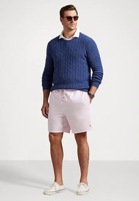 Blå kabelstrikket genser, hvit skjorte med krage under, lyserosa shorts med tynne striper, hvite joggesko og solbriller.