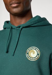 Groene hoodie met een trekkoord en een cirkelvormig grafisch logo met een gestileerde golf en tekst die milieuthema's belicht.
