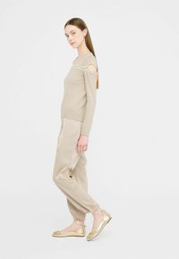 Beige Strickpullover mit ausgeschnittenen Schultern, kombiniert mit hellbeigen Hosen. Die Schuhe sind metallisch goldene Ballerinas mit dekorativen Akzenten.