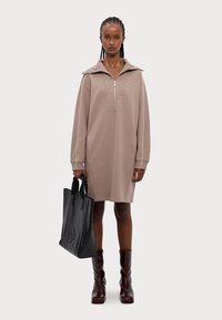 Abito felpa beige oversize con chiusura a zip e ricamo frontale. Abbinato a una borsa tote nera e stivali al ginocchio lucidi bordeaux.