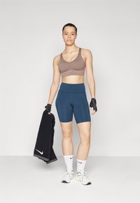 Brun sport-BH, marineblå sykkelshorts, svart håndkle, hvite sko og sokker. Modellen har kort hår og har på seg hansker.