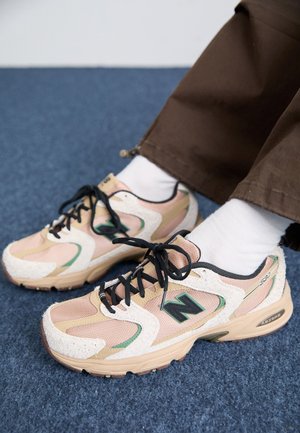 Person trägt beige und hellrosa New Balance Sneaker mit schwarzen Schnürsenkeln, weißen Socken und brauner Hose auf blauem Teppichboden.
