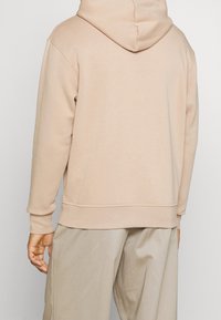 Felpa beige con cappuccio e vestibilità comoda, realizzata in tessuto liscio, caratterizzata da un cappuccio ampio e polsini a costine, su uno sfondo neutro.