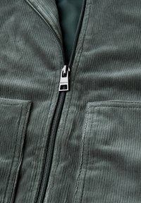 Green corduroy jacket featuring a silver zipper with a pull tab, two front pockets, and textured fabric in a vertical rib pattern.