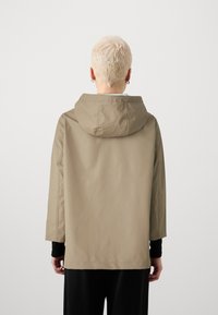 Chaqueta impermeable beige con capucha, con mangas largas y un corte relajado. Textura suave con costuras mínimas y un corte recto.