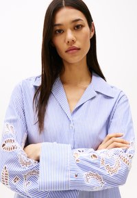 Camicia a righe blu e bianche con colletto e maniche lunghe, caratterizzata da dettagli in pizzo bianco sui polsini.
