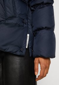 Main reposant à côté d'une doudoune bleu marine avec boutons-pression et étiquette "Marc O'Polo", portée sur un pantalon en cuir noir.