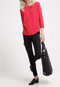 Blusa roja de manga larga con cuello redondeado, combinada con pantalones negros ajustados y mocasines negros. Se sostiene un bolso negro en una mano.