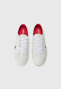 Baskets blanches avec une tige lisse, une semelle en caoutchouc texturé, un intérieur rouge, des lacets et un petit logo en forme de cœur rouge sur le côté.