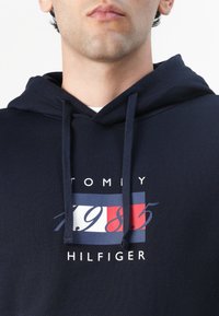 Sweat à capuche bleu marine avec une poche kangourou, orné d'un imprimé graphique audacieux en rouge, blanc et bleu, affichant "TOMMY 1985 HILFIGER."