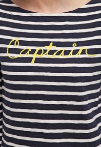 Marineblau-weiß gestreiftes Hemd mit rundem Ausschnitt; mit gelber gestickter Aufschrift "Captain" über der Brust.