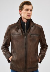 Veste en cuir marron avec col montant, zip avant et deux poches poitrine. Finition texturée avec de subtils détails de couture.