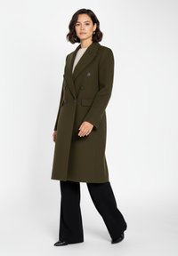 Olivgrön dubbelknäppt kappa med texturerat tyg, slagkrage och svarta knappar, kombinerad med svarta byxor och spetsiga skor.