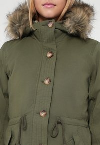 Olive roheline parka jakk kunstkarusnahast kapuutsiga, nööpide eesmine kinnitamine, reguleeritav vöökoht ning nähtavad õmblusdetailid. Sile tekstuur.