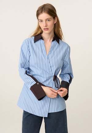 Vrouw draagt een blauw gestreept wikkelshirt met donkerbruine kraag en manchetten, strik het zijlint, gecombineerd met donkerblauwe broek.