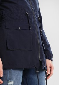 Granatowa kurtka z zamkiem błyskawicznym, regulowanymi sznureczkami i dwoma dużymi kieszeniami z przodu. Gładka tekstura i lekki materiał.