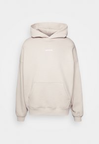 Sweat à capuche beige oversize avec poche kangourou à l'avant et petit texte blanc "WRSTBHVR" centré sur la poitrine.