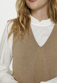 Hnědý pletený svetr s výstřihem do V, zdobený texturovaným vzorem a žebrovanými okraji, vrstvený přes bílou blůzu s rýškovým límcem.