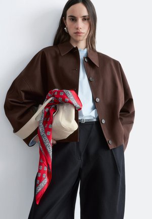 Femme portant une veste marron boutonnée et un pantalon noir, tenant un sac beige orné d'une écharpe rouge à motifs sur un fond uni.