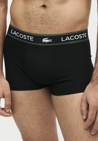 Homme portant un boxer noir Lacoste avec ceinture et logo bordé de vert, montrant le bas du torse et le haut des cuisses sur fond uni.