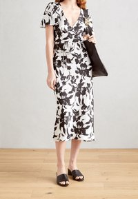 Robe portefeuille fleurie blanche avec des fleurs noires, manches courtes volantées et ceinture à nouer. Associée à des mules à talons blocs ouverts noires.