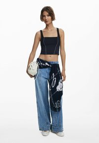 Černý crop top s čtvercovým výstřihem, modré široké kalhoty, a tmavý vzorovaný šátek uvázaný v pase. Bílá kulatá kabelka.