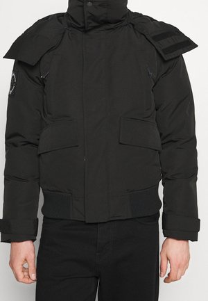 Schwarze gesteppte Jacke mit hohem Kragen, Klappentaschen, verstellbaren Manschetten und einem Aufnäher am oberen linken Ärmel, getragen über einer schwarzen Hose.