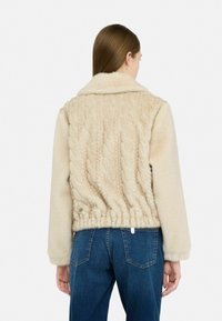 Giacca in finta pelliccia beige con una trama a maglia cable. Design cropped, con colletto e orlo elasticizzato. Abbinata a jeans in denim blu.