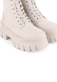 Botas altas color beige con cordones, con una suela de goma texturizada, que presenta una línea de acento naranja y un upper de cuero suave. Diseño robusto con punta redondeada.