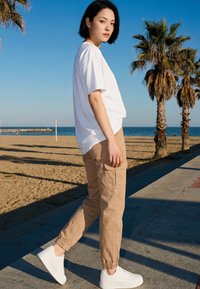 Junge Frau in weißem T-Shirt, beiger Hose und weißen Sneakers läuft auf einem Strandweg mit Palmen und Meer im Hintergrund.