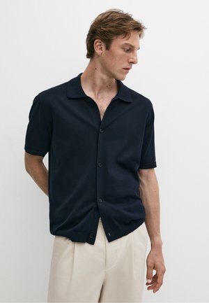 Mann trägt ein schwarzes kurzärmeliges Hemd mit Knöpfen und hellbeige Hose, schaut nach unten und zur Seite vor weißem Hintergrund.