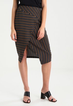 Femme portant une jupe midi noire avec des rayures diagonales marron et blanches, fendue à l'avant, associée à des sandales noires à talons blocs ouverts.