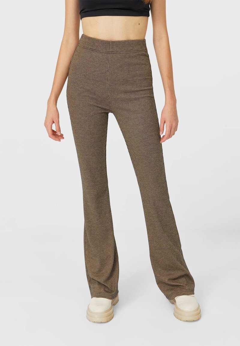 Pantalons évasés à motif marron avec une taille haute, présentant un design en pied-de-poule. Confectionnés en tissu texturé, associés à des chaussures beiges.