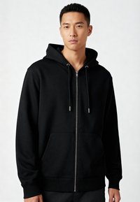 Giovane che indossa un semplice sweat hoodie nero con zip e cordini, in piedi contro uno sfondo neutro chiaro.