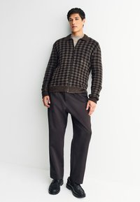 Veste zippée à carreaux marron avec un col côtelé, associée à un pantalon foncé et des chaussures noires. Texture lisse, teintes contrastées foncées et claires.