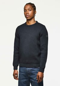 Mann mit geflochtenem Haar steht mit Blick nach vorne, trägt einen schwarzen Rippstrickpullover und blaue Jeans vor einem schlichten hellen Hintergrund.