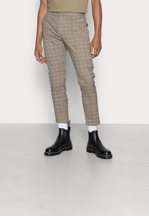 Osoba nosząca beżowe, w kratę, dopasowane spodnie, białe skarpetki i czarne skórzane botki Chelsea na szarej podłodze na jasnym, jednolitym tle.