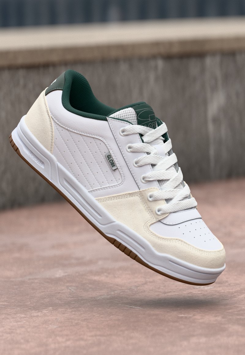Adidași albi cu accente verzi, din suede texturat și piele, șireturi plate și talpă din cauciuc. Prezintă perforații și un logo de marcă.