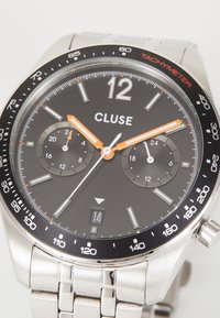 Ανοξείδωτο ρολόι με μαύρο καντράν, ασημένιους δείκτες ωρών και πορτοκαλί λεπτομέρειες. Διαθέτει υποκαντράν χρονογράφου, στεφάνι ταχυμέτρου και παράθυρο ημερομηνίας.