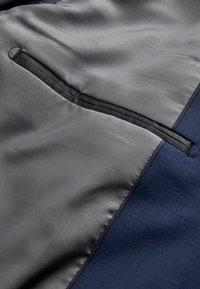 Marineblauwe stof met een gladde, matte textuur heeft een grijze satijnen zak met een zwarte afwerking en een klein, cirkelvormig stiksel detail.